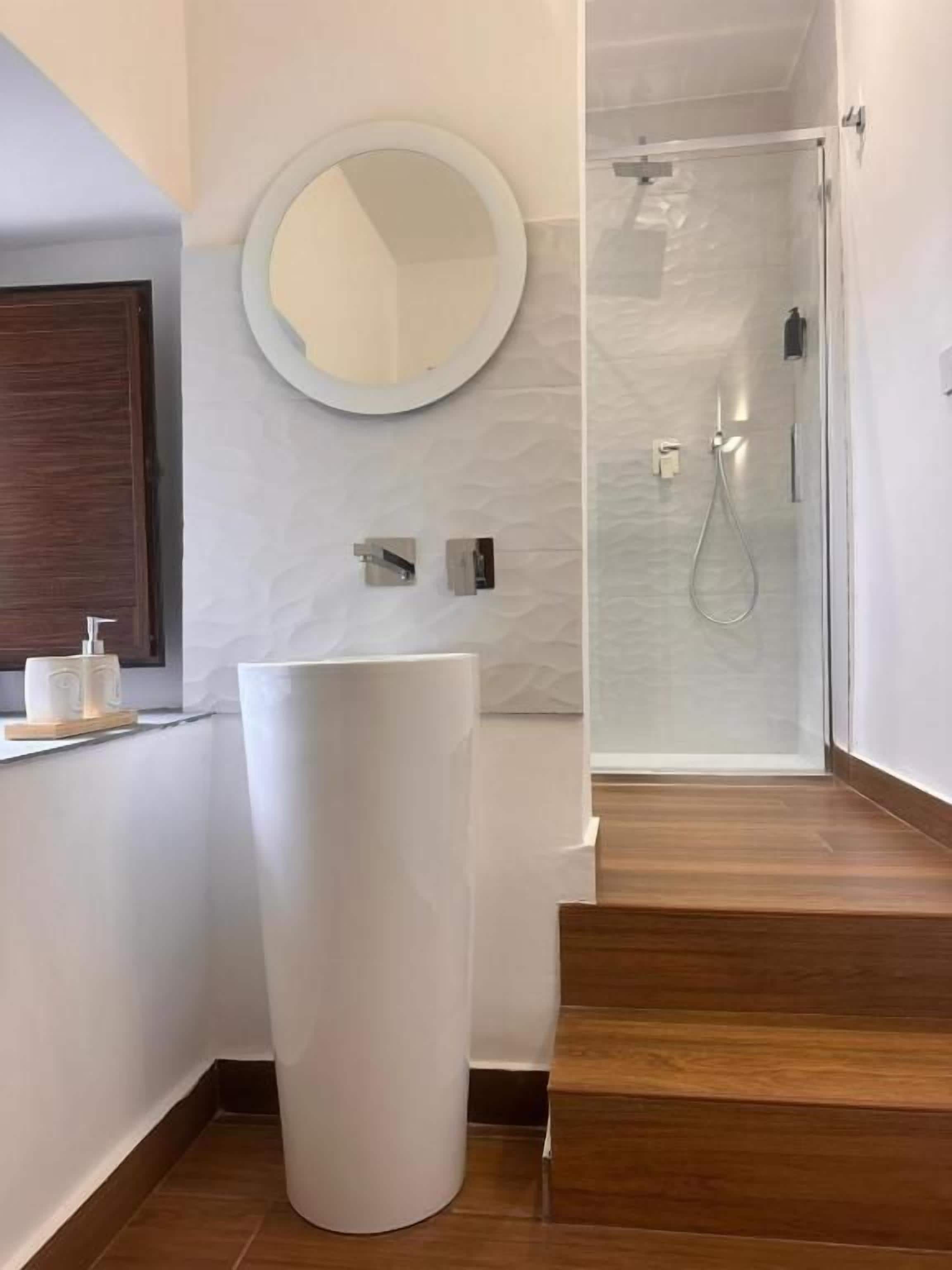 Une salle de bain moderne avec un grand miroir rond, un lavabo sur pied blanc et une douche à l’italienne.
