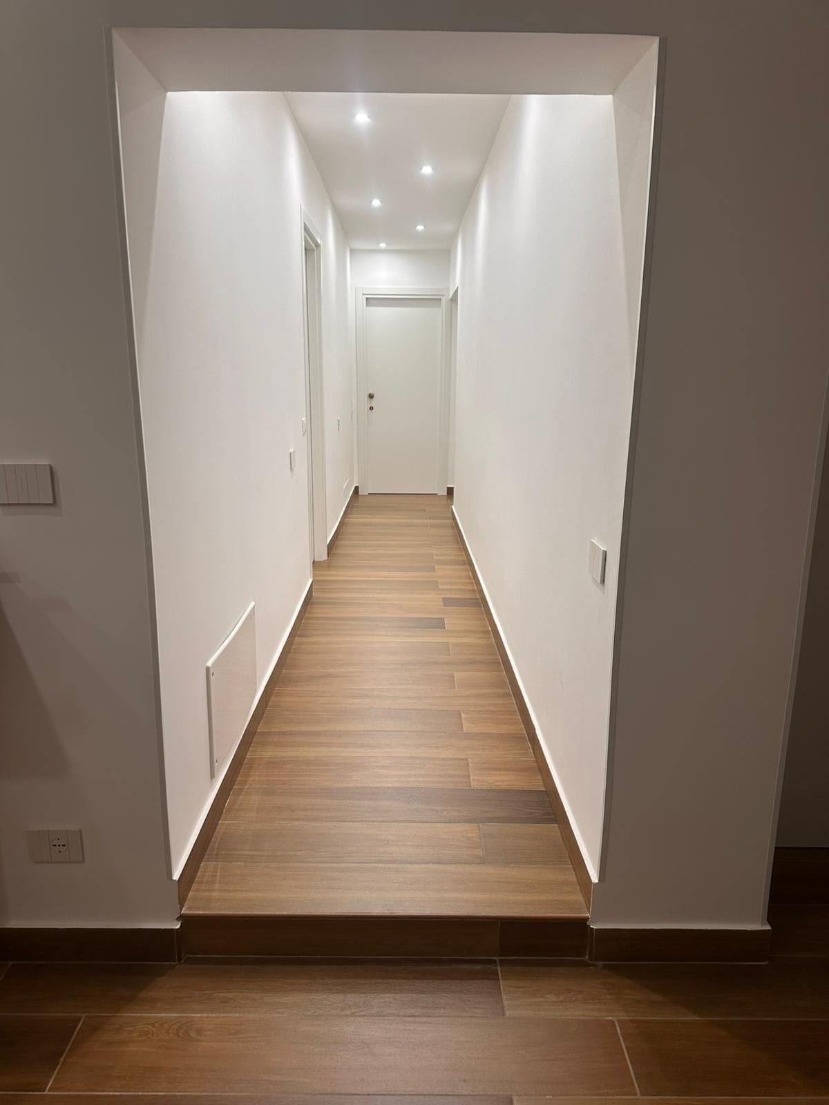 Un couloir étroit et bien éclairé, doté d’un plancher en bois et de murs blancs.