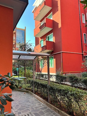 A courtyard with a covered seating area, a red building with balconies, and a clear blue sky.