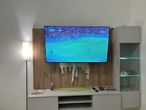 A television mounted on a wall with a wooden panel backdrop, a remote control, and a decorative vase on a white surface below.