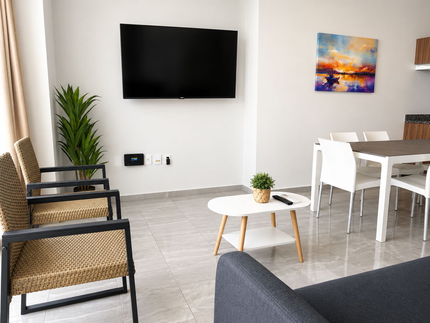 Un salón moderno con televisor de pantalla plana, una zona de comedor con sillas blancas, una pequeña mesa redonda con una planta, y una gran planta en maceta.