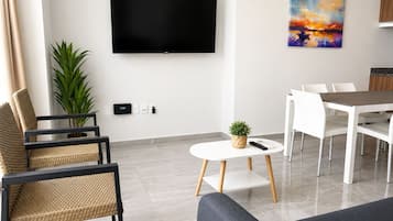 Un salón moderno con televisor de pantalla plana, una zona de comedor con sillas blancas, una pequeña mesa redonda con una planta, y una gran planta en maceta.