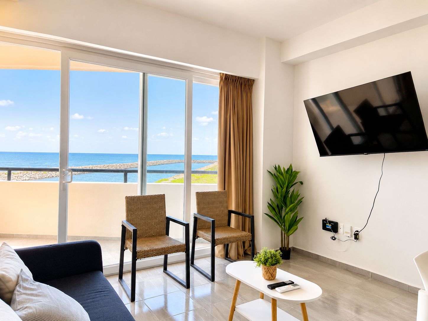 Una sala moderna con un ventanal grande, dos sillones de mimbre, un sofá, una mesita con una planta y un televisor montado en la pared.