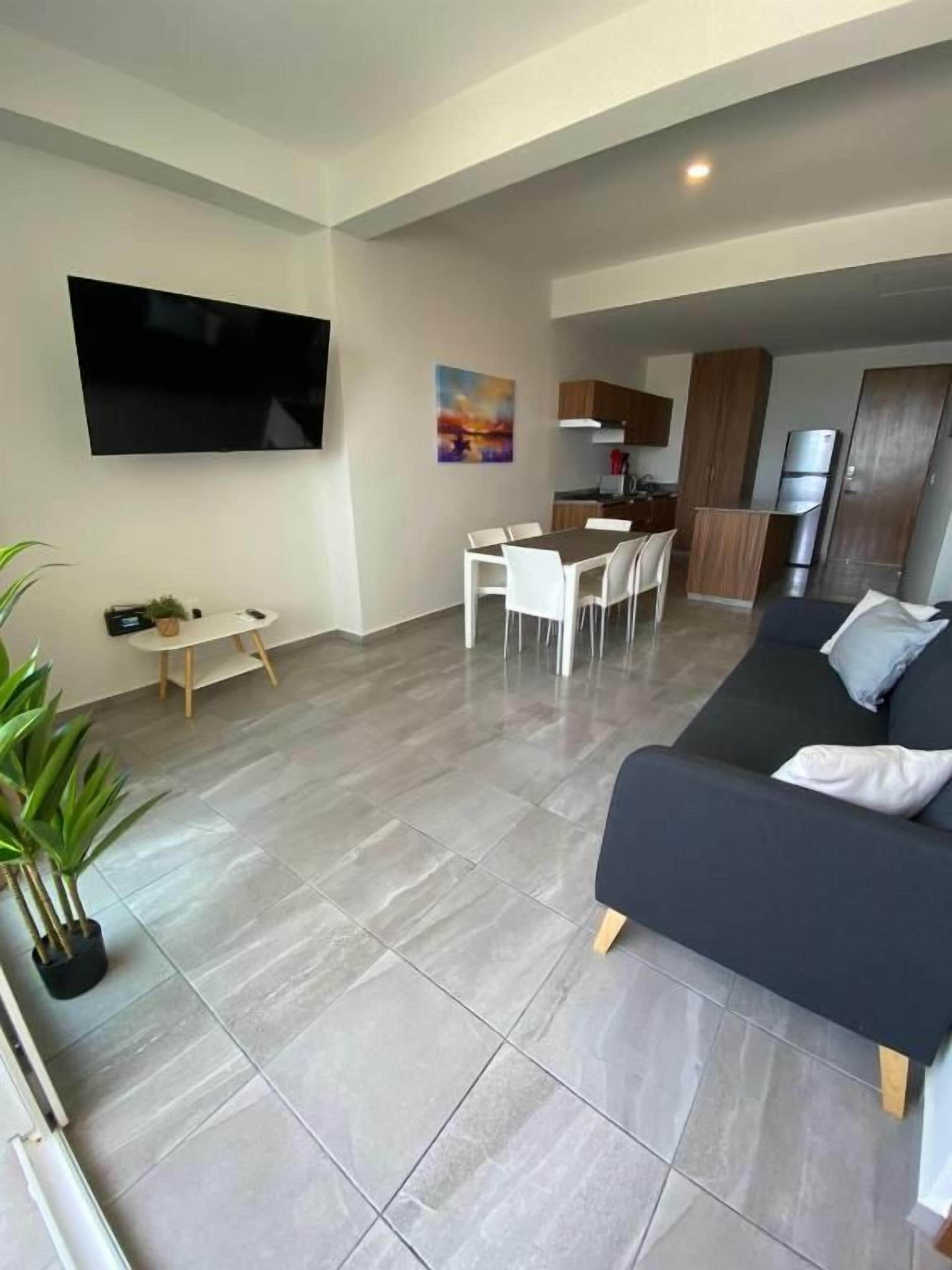 Una sala moderna con un televisor de pantalla plana, un sofá, una mesa de comedor con sillas y una cocina al fondo.
