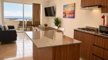 A modern kitchen with a central island, a dining area with chairs, a TV mounted on the wall, and a view of the ocean through the sliding glass door.