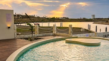 A swimming pool with a view of a sunset over a body of water, surrounded by a wooden deck and a low wall.