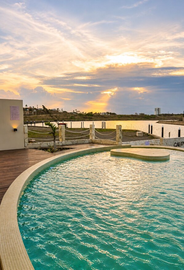 Une piscine avec vue sur un coucher de soleil se reflétant sur une étendue d’eau, entourée d’une terrasse en bois et d’un petit muret.
