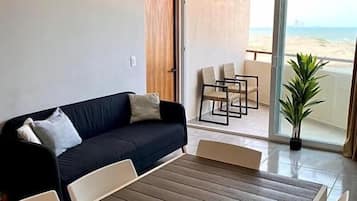 Habitació amb sofà, taula de menjador i vista a la platja a través d'una porta de vidre.