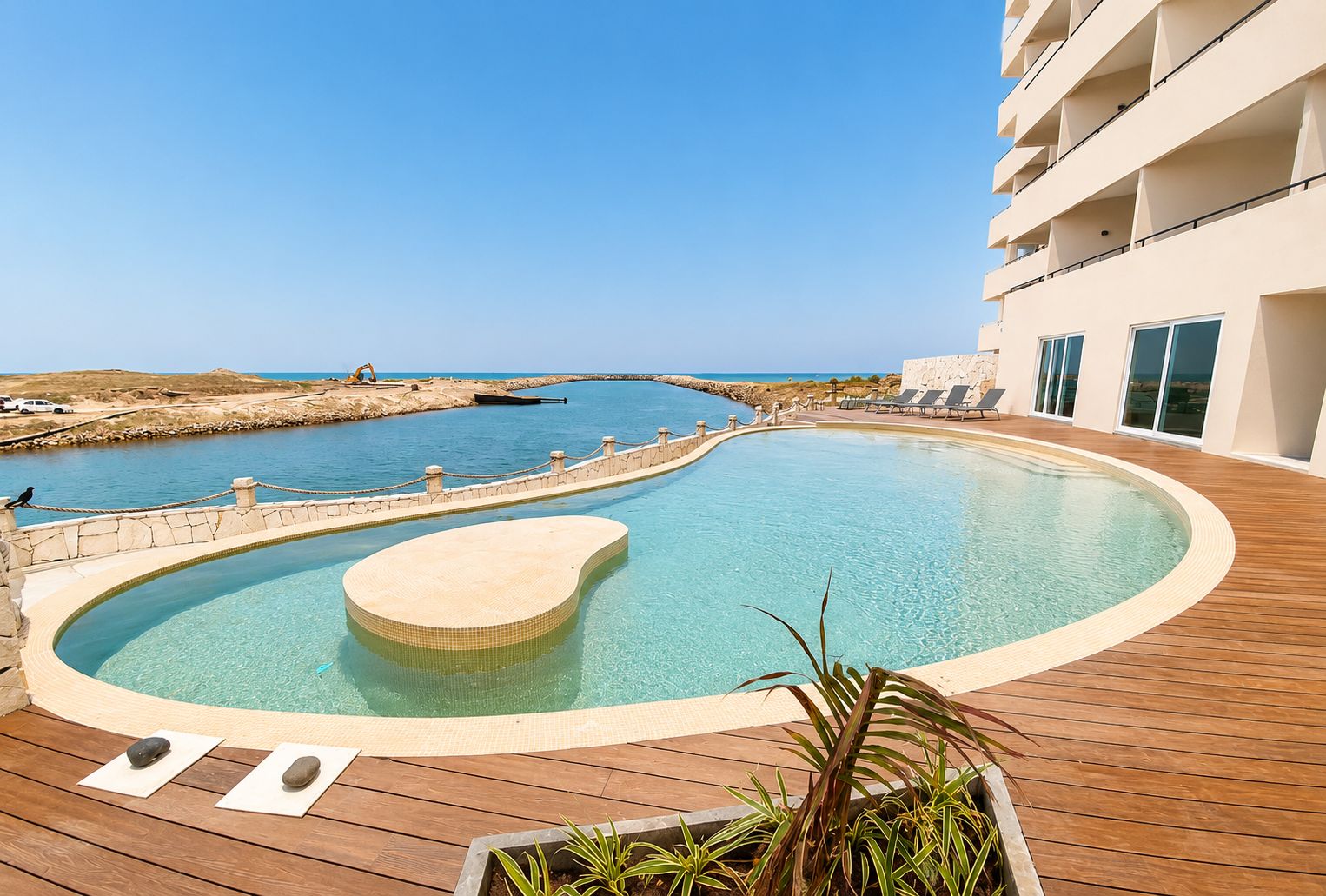 Una piscina con plataforma de madera, pared de piedra y vista al mar.