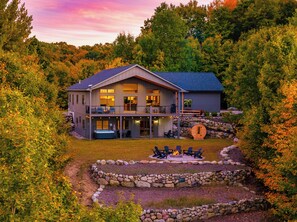 A two-story house with a balcony, a hot tub, and a stone fire pit surrounded by a stone wall. - Long-Range Lake Views | Sauna, Plunge, Hot Tub (Bellaire)