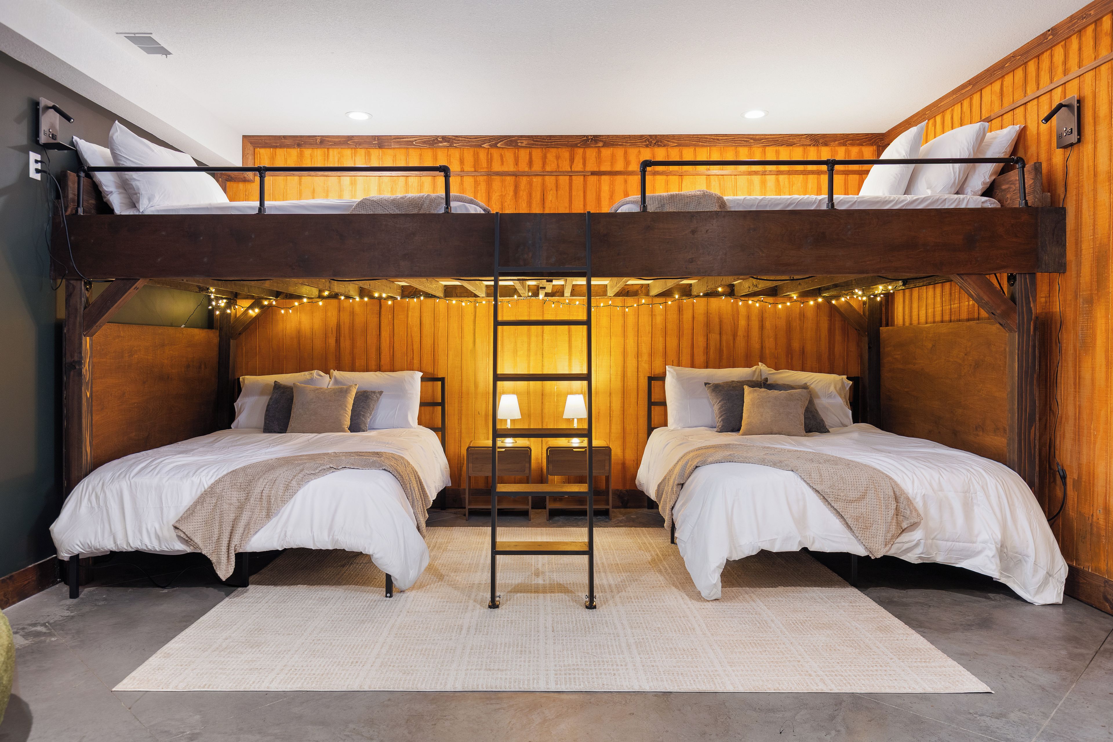 A bunk bed room with wooden walls, a central ladder, and string lights.