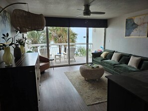 A living area with a sofa, a dining table, and a balcony with a view of palm trees. - Rising Tides Properties Condo 204 (Murrells Inlet)