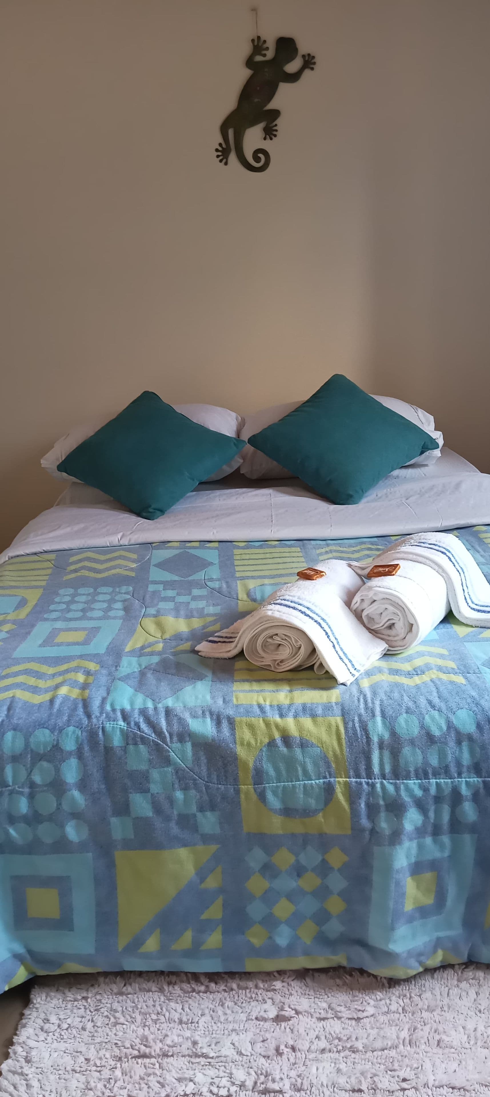 A bed with a patterned bedspread, two teal pillows, and a rolled white towel.