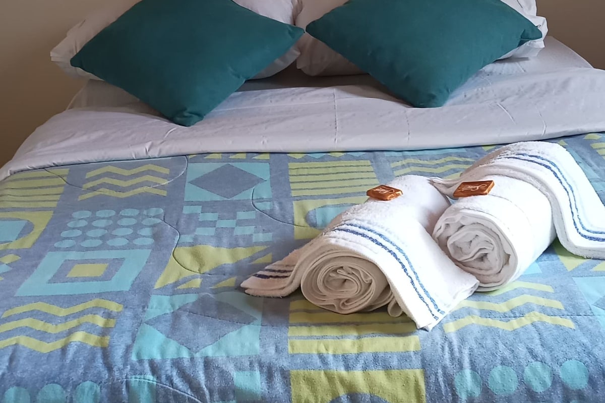 Una cama con una colcha estampada, dos almohadas color turquesa y una toalla blanca enrollada.