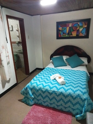 Un dormitorio con cama, almohadas, una colcha, un cuadro en la pared y una puerta que da a otra habitación.