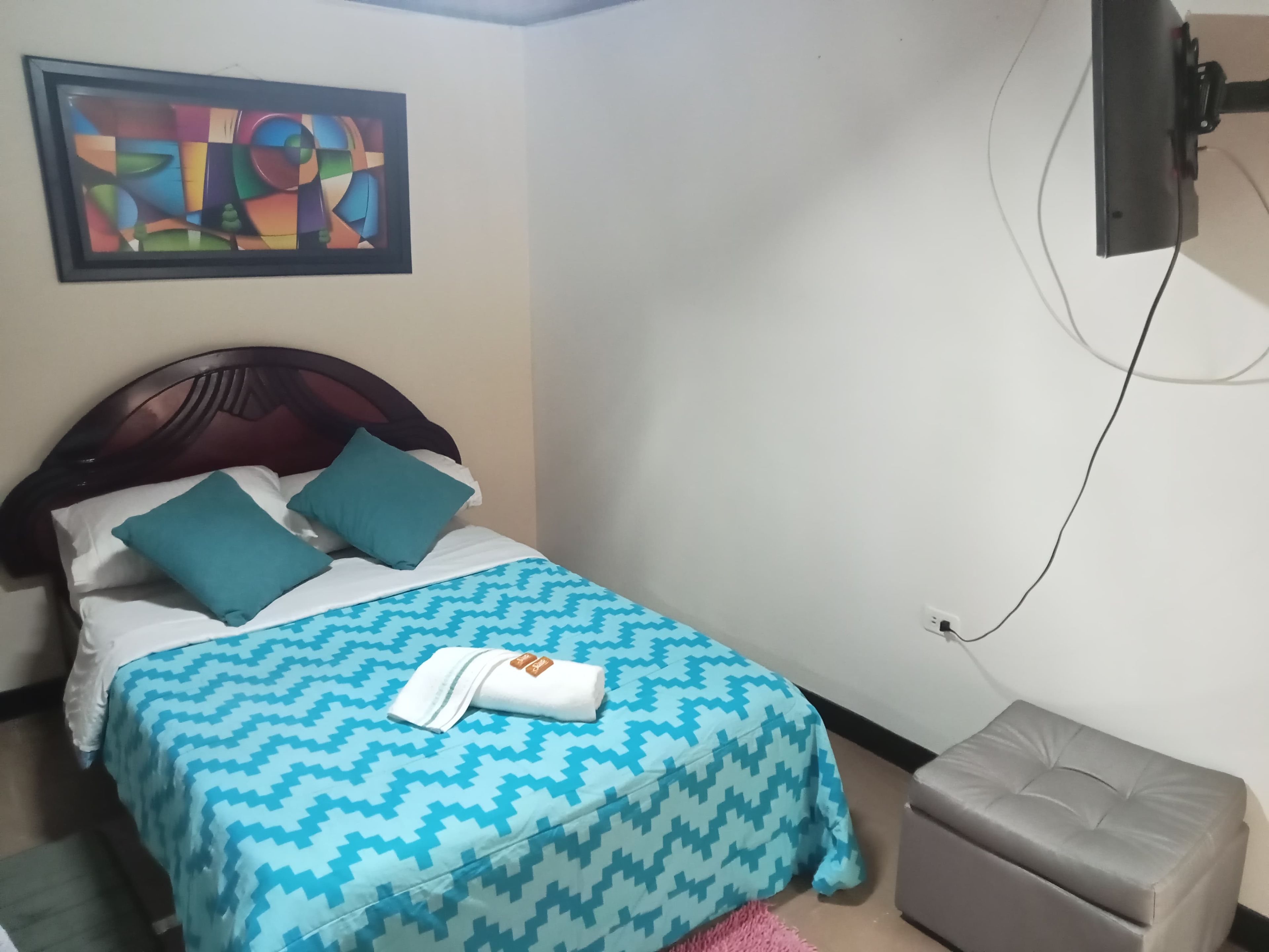 A bed with a patterned bedspread, two pillows, a folded towel, a television mounted on the wall, and a small ottoman.