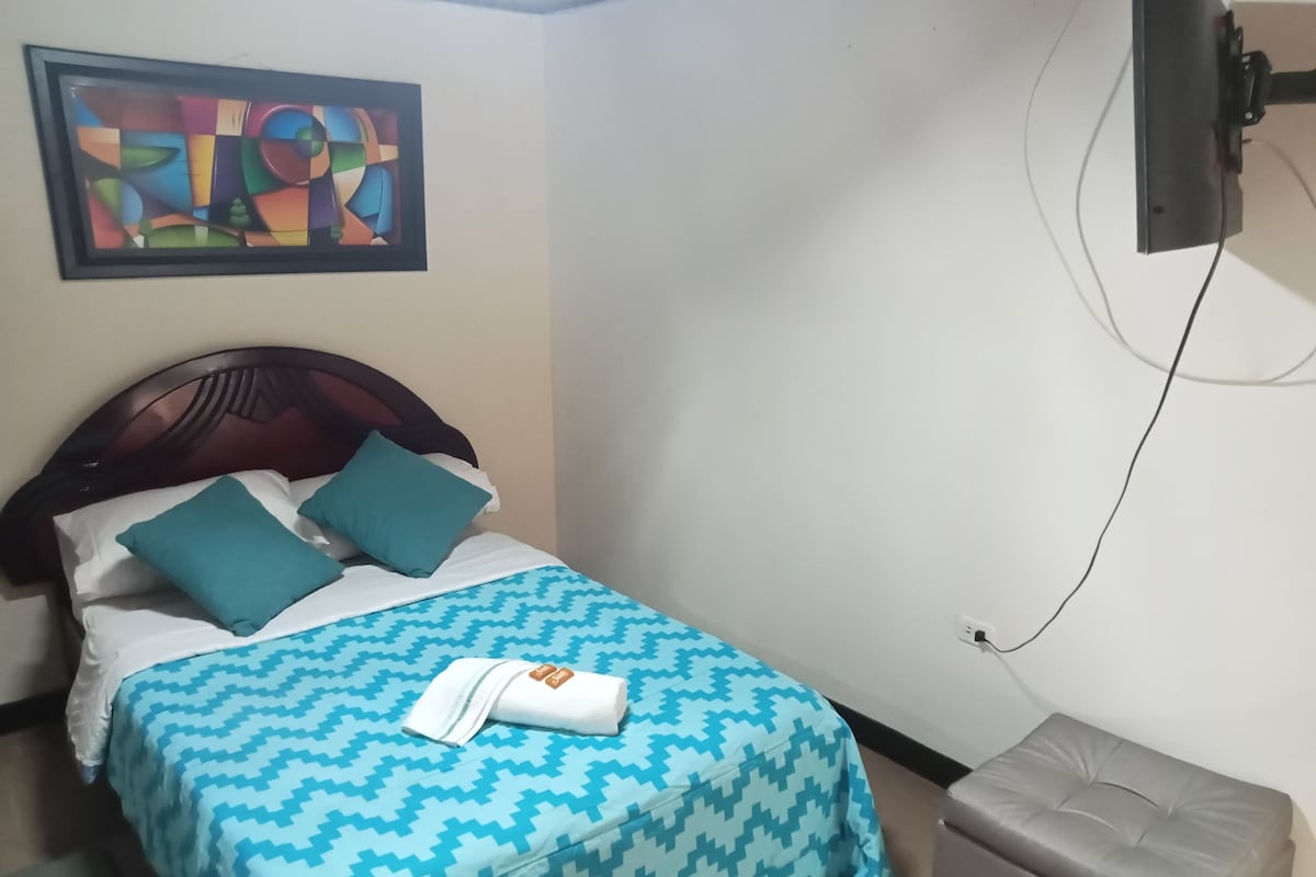 Una cama con colcha estampada, dos almohadas, una toalla doblada, un televisor montado en la pared y un pequeño otoman.