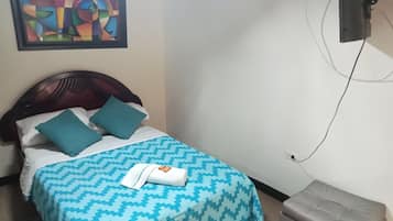 A bed with a patterned bedspread, two pillows, a folded towel, a television mounted on the wall, and a small ottoman.