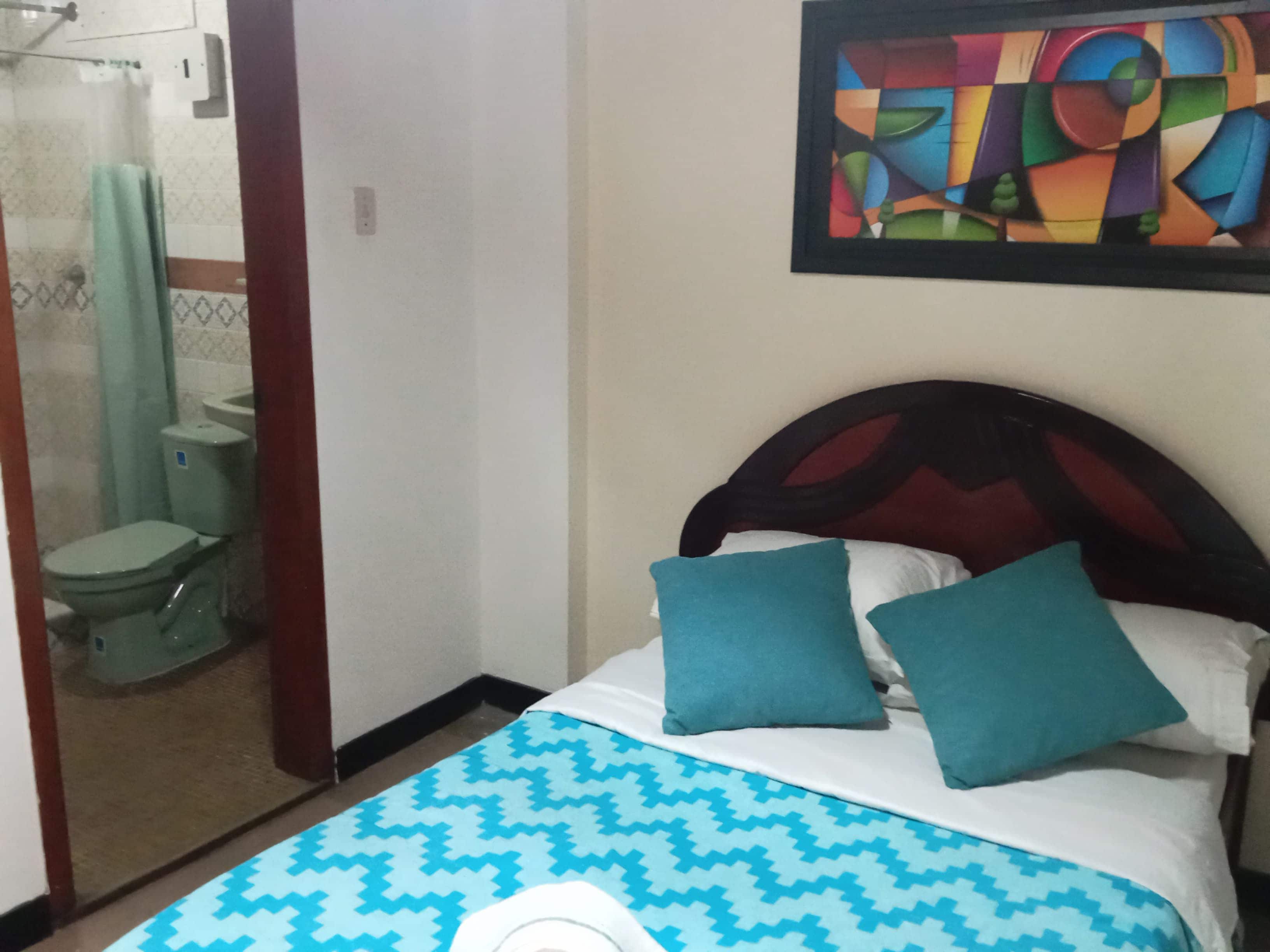A bedroom with a bed, two pillows, a wall painting, and a bathroom visible through an open door.