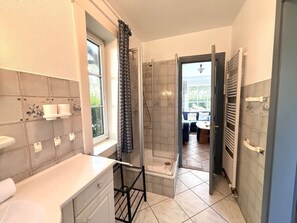 A bathroom with a shower, a toilet, and a sink. There is a window with a curtain, and a door leading to a balcony. - "oldsum 2, House 94" (Oldsum)