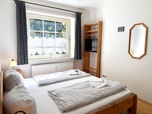 A bedroom with two beds, a window with curtains, a radiator, a wooden shelf with a TV, and a wall-mounted mirror. - "oldsum 2, House 94" (Oldsum)