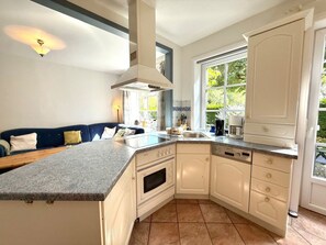 A kitchen with a built-in island, white cabinetry, a window with a view, and a blue sofa in the adjacent living area. - "oldsum 2, House 94" (Oldsum)