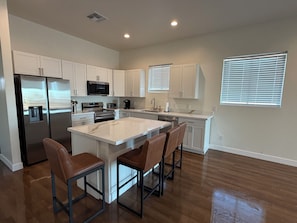 A modern kitchen with white cabinetry, stainless steel appliances, a central island with bar stools, and a large window with blinds. - Spacious 4-bedroom new construction house in fantastic Surprise with AC (Surprise)