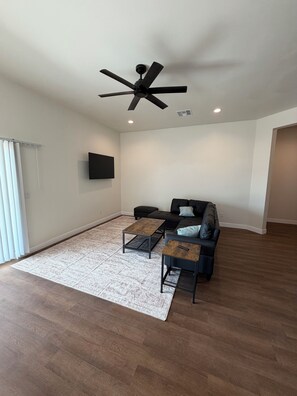 A spacious living room with a ceiling fan, a black sofa, two coffee tables, and a large window with blinds. - Spacious 4-bedroom new construction house in fantastic Surprise with AC (Surprise)