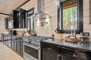 A modern kitchen with stainless steel appliances, wooden cabinetry, and a large window offering a view of trees. - Sleeps 20 w/Playset·Arcade + Outdoor Pool Table (Broken Bow)