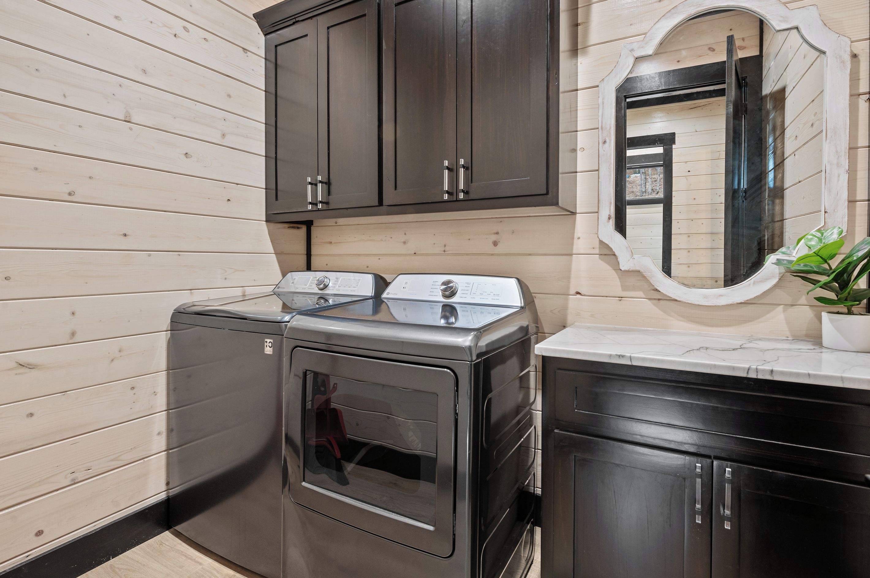A compact laundry room with a front-loading washer and dryer, wooden panel walls, and a mirror above the sink.