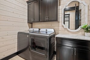 A compact laundry room with a front-loading washer and dryer, wooden panel walls, and a mirror above the sink. - Sleeps 20 w/Playset·Arcade + Outdoor Pool Table (Broken Bow)