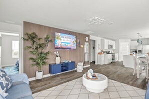 Uma sala de estar moderna com televisão de tela plana, um sofá azul, uma mesa de centro redonda e uma área de cozinha com armários e eletrodomésticos brancos.