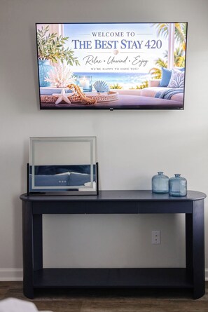 Ein an der Wand befestigter Fernseher zeigt eine Willkommensbotschaft für 'The Best Stay 420' und bietet Blick auf die Küste. Darunter befindet sich eine dunkle Konsole mit einem gerahmten Bild und zwei Glasvasen.