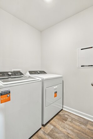 A laundry room with two white washing machines and a dryer, a white wall, and a wooden floor. - The Best Stay 420 - Cozy 4BR Home Near Savannah (Port Wentworth)