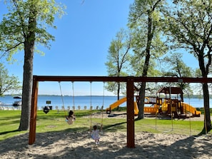A playground with swings and a slide, set against a backdrop of trees and a body of water.