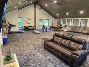 A spacious indoor area with a brown leather sofa, a pool table, a ping pong table, a treadmill, and a dining table with chairs.