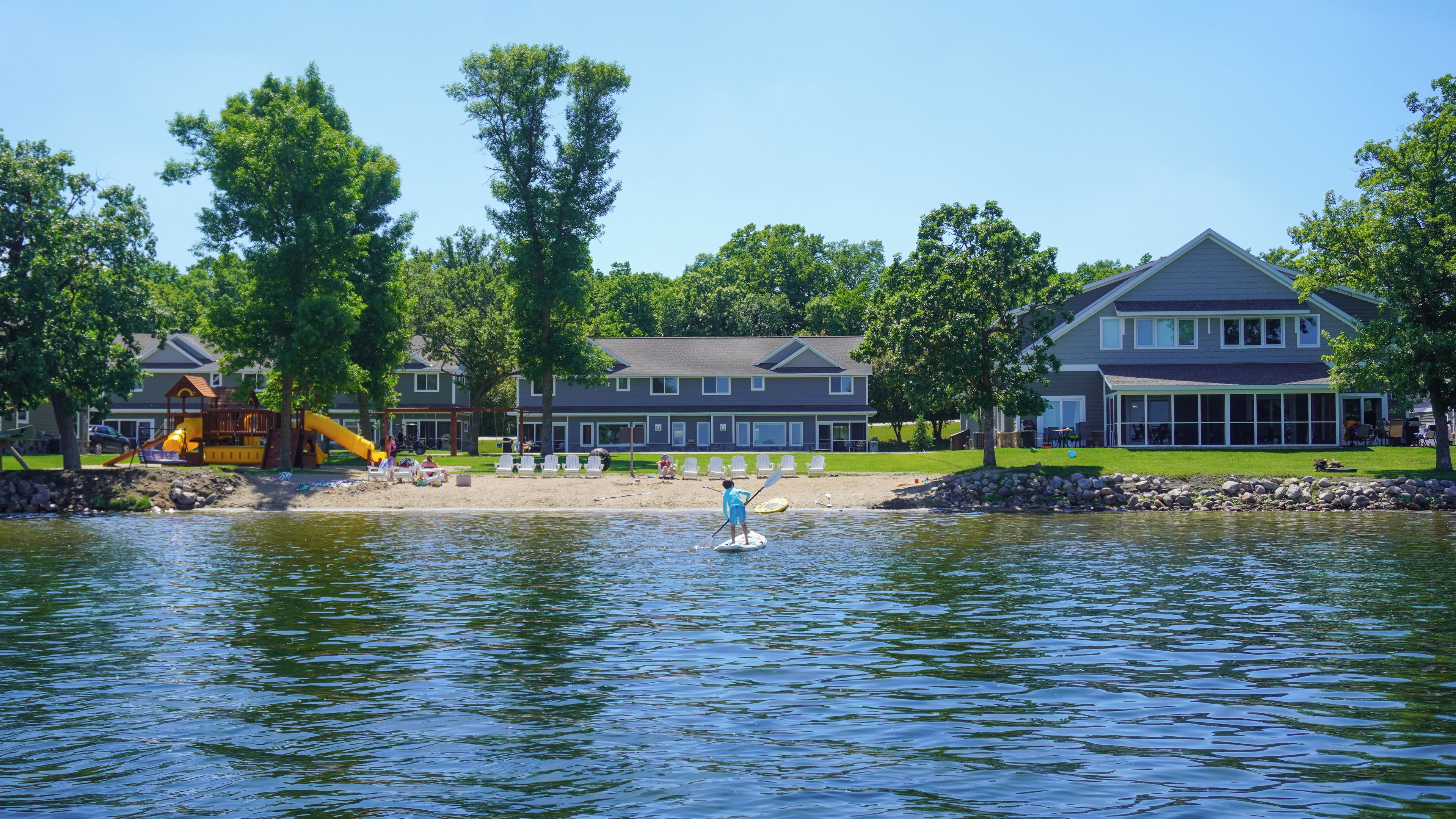 A lakeside property with a dock, a playground, and a large house with a covered patio.
