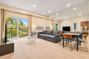 A modern living room with a sofa, dining table, and chairs, a TV, and a balcony with a view of greenery. - Renovated Condo Near Omni La Costa Resort & Golf! (Carlsbad)