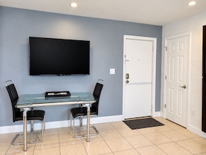 A modern living room with a glass-top table, two black chairs, a flat-screen TV mounted on the wall, and a white door. - Coastal Modern One-Bedroom Apartment (St. Pete Beach)