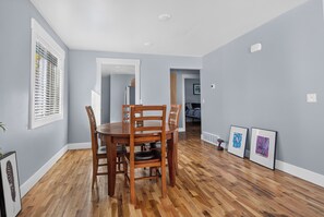 Un coin repas avec une table et des chaises en bois, une fenêtre avec des stores et un couloir menant à une chambre.