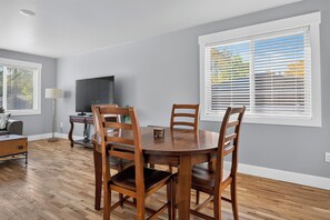 A dining area with a wooden table and chairs, a television on a stand, and a window with blinds. - Newly Remodeled 4 Bedroom Townhome for 8 Guests (Billings)