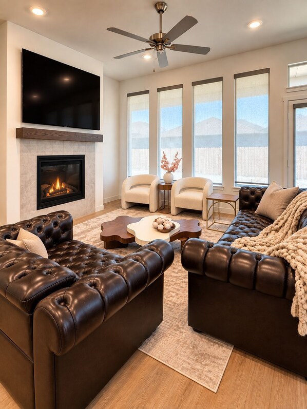 A modern living room with leather sofas, a fireplace, a ceiling fan, and large windows.
