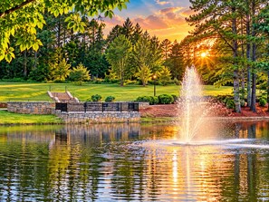 A serene lakeside scene with a fountain, stone steps, and a grassy area with trees. - ATL Lakeview Haven - Private Suite & Deck (Decatur)