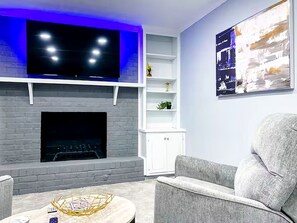 A modern living room with a flat-screen TV mounted above a fireplace, a white shelving unit, and a grey sofa. - ATL Lakeview Haven - Private Suite & Deck (Decatur)