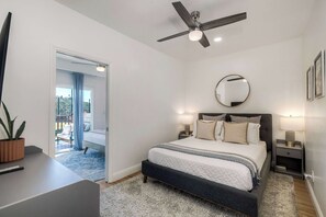 Ein modernes Schlafzimmer mit Bett, Nachttischlampen, Deckenventilator, rundem Spiegel und Blick ins Freie.