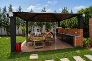 En udendørs patio med et overdækket spiseområde, grillstation og skraldespand.