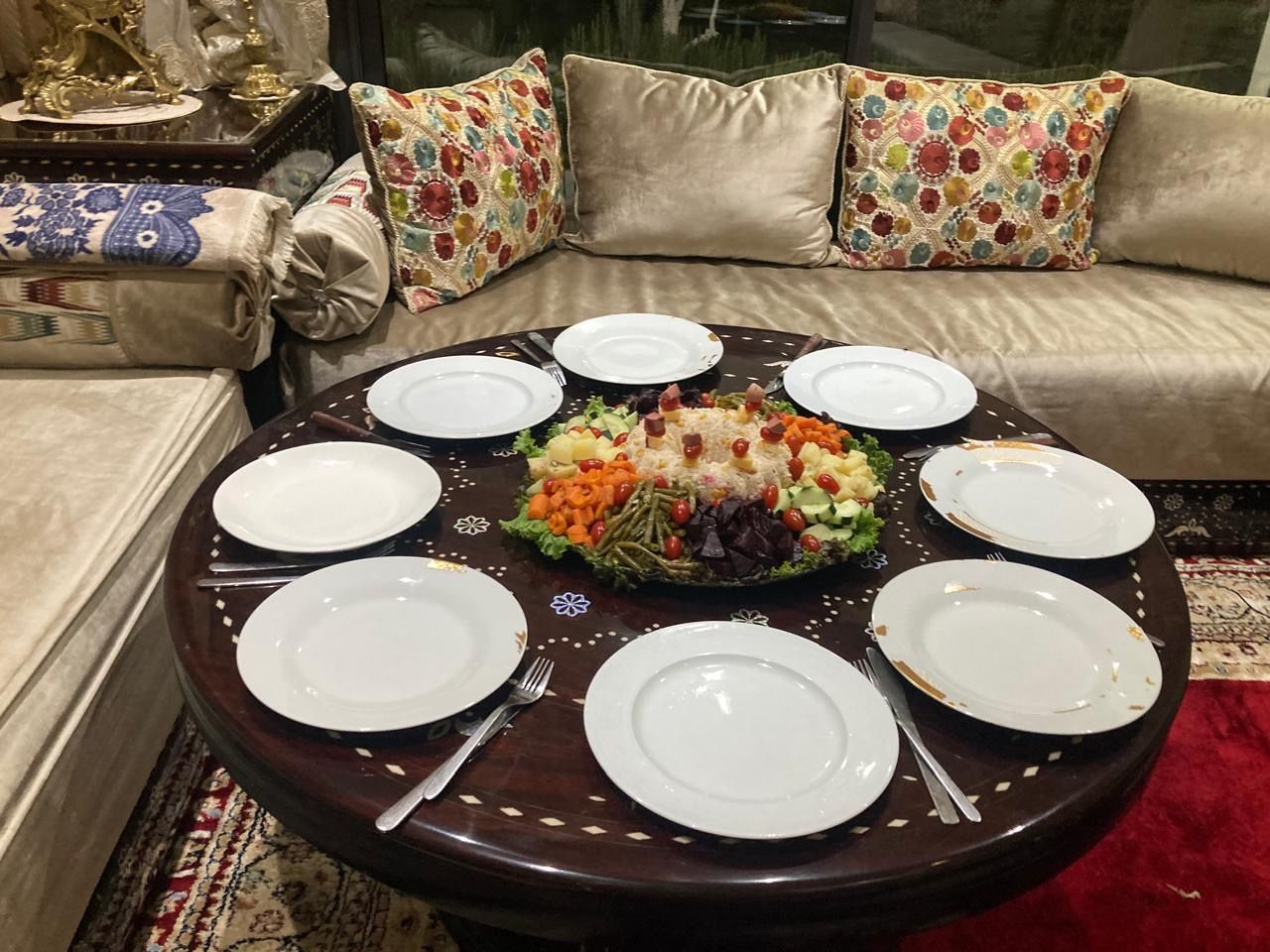 A round dining table with white plates and cutlery, a central vegetable platter, and a sofa with patterned cushions in the background.