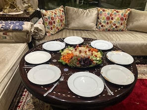 A round dining table with white plates and cutlery, a central vegetable platter, and a sofa with patterned cushions in the background.