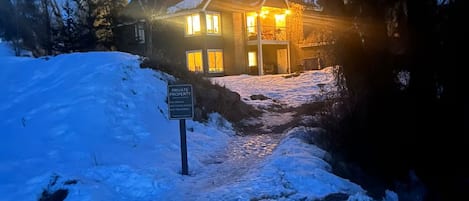 Casa de dos pisos con puerta y ventanas iluminadas, rodeada de nieve en el suelo y árboles.