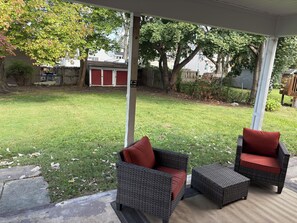 Een overdekt terras met rieten meubels en een rode kussen, een gazon en een houten omheining op de achtergrond.